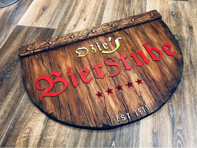 Dřevěná cedule ve stylu starého dřeva s nápisem „Ozie’s BierStube“ z nalepených písmen ve staroněmeckém písmu